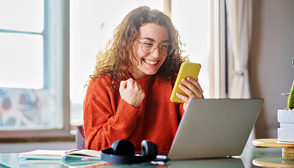 Frau sitzt am Laptop und freut sich, w&auml;hrend sie auf ihr Smartphone schaut.